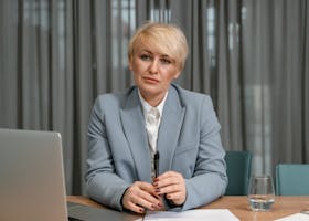 A confident businesswoman in a blue suit sits at her desk with a laptop, pen, and papers in an office setting.